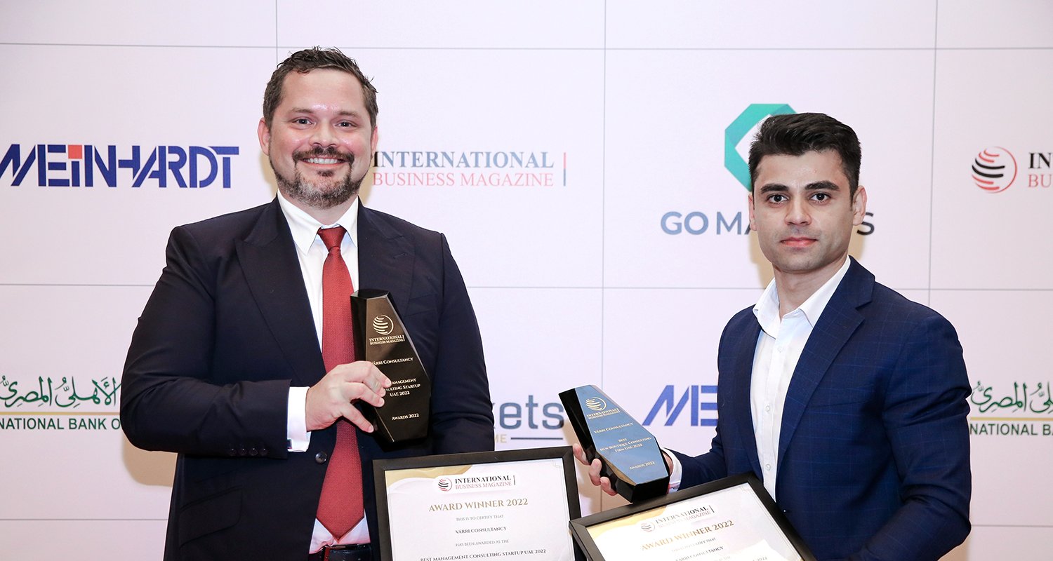 Two individuals holding trophies and framed certificates for “Best New Boutique Consulting Firm UAE 2022” and “Best Management Consulting Startup UAE 2022”, awarded to Várri Consultancy at the International Business Magazine Awards 2022 ceremony, standing in front of a sponsor-branded backdrop.
