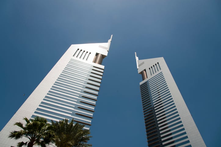 The Emirates Towers in Dubai, UAE, shot from below with the sun reflecting in the building and a clear blue sky behind. A palm tree in the forefront.