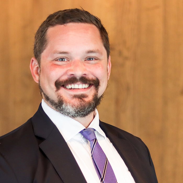 A close-up of Johnny Kollin in business attire, smiling, a wooden wall behind him.